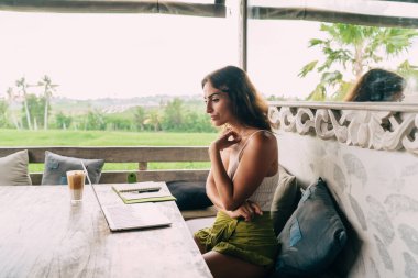 Süslü yaz kıyafetleri içinde, tropikal tatil beldesindeki terasta otururken dizüstü bilgisayarından nahoş haberler okuyan düşünceli, genç İspanyol kadının yan görüntüsü.