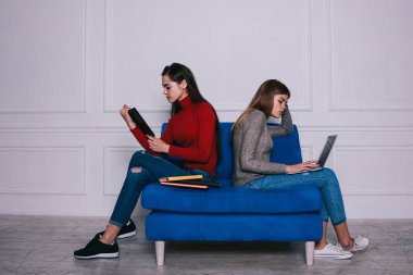 Gömülmüş kadınlar, günlük kazaklar ve kot pantolonlar içinde dizüstü bilgisayar ve kitaplarla ödevlerini yaparken mavi koçun üzerinde oturuyorlar.