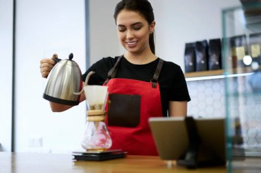 Gülümseyen yetenekli bayan barista kahve hazırlarken cam filtre garı ve çaydanlıktan sıcak su kullanıyor.