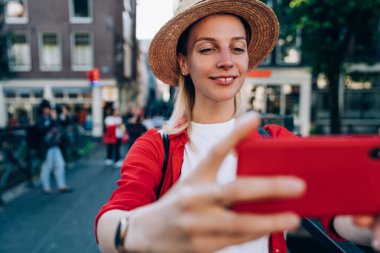 İyimser bayan gezgin hasır şapka ve sırt çantalı sıradan kıyafetler giyerek Amsterdam 'ı gezerken kendi portresini çekerken gülümsüyor.
