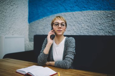 Gündelik giysiler içinde, açık defterle masada oturan ve cep telefonuyla konuşan serbest çalışan genç bir kadının yumuşak odağı.