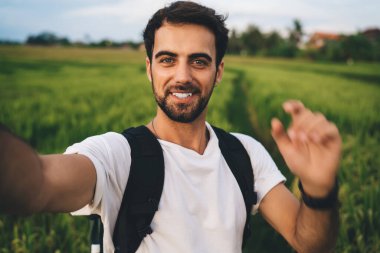 Beyaz tişörtlü, yeşil alanda sırt çantalı ve yaz tatilinde selfie çekerken kameraya gülümseyen yakışıklı adam.