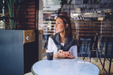 Kafe terasında oturan gülümseyen beyaz kadın bloglar için 4G internete bağlı akıllı telefon kullanıyor.