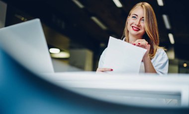 Olumlu bir kadının, modern çalışma ortamında masa başında otururken kağıtları kontrol ederken ve uzaktan bilgisayar başında çalışırken yan gözle baktığı düşük açılı bir bakış açısı.