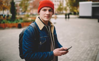 Sırt çantalı yakışıklı erkek şehir caddesinde yürürken cep telefonundan mesaj yazan kameraya bakıyor.