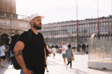 Haftasonu güneşli bir günde şehir merkezinde tarihi sarayın yanında duran sırt çantalı ve şapkalı olumlu bir erkek.