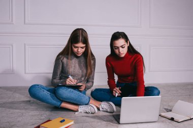 Genç, odaklanmış kız öğrenciler dizüstü bilgisayarla yeni bir proje üzerinde çalışıyorlar ve yere oturup bacak bacak bacak üstüne atarak notlar alıyorlar.