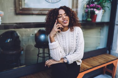 Kol saati takılı gülümseyen Afro-Amerikalı kadın ahşap bankta oturuyor ve sokakta dinlenirken akıllı telefondan konuşuyor.