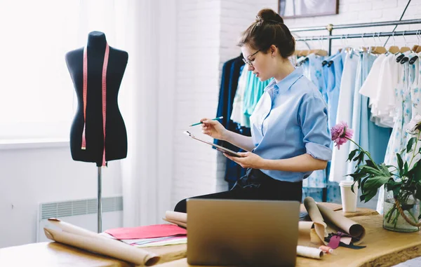 Gözlüklü ve gömlekli ciddi bir terzinin modern atölyede moda projesinin detaylarını düşünmesi ve not alması.