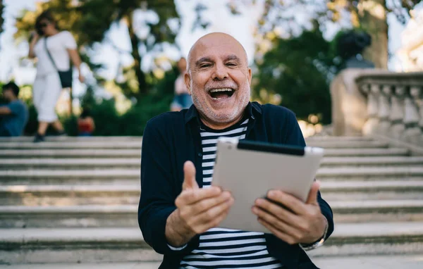 Half length portrait of cheerful senior traveller with modern touch pad gadget laughing at camera during free time in city