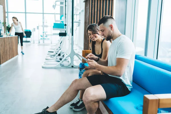 Personal trainer sitting on couch and instructing sporty woman while browsing tablet in modern gym with equipment during break from workout