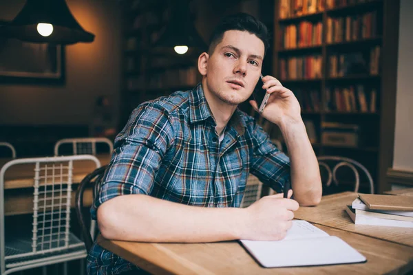 Thoughtful man in casual clothes sitting at table in library and writing in notebook while having phone conversation and pensively looking away
