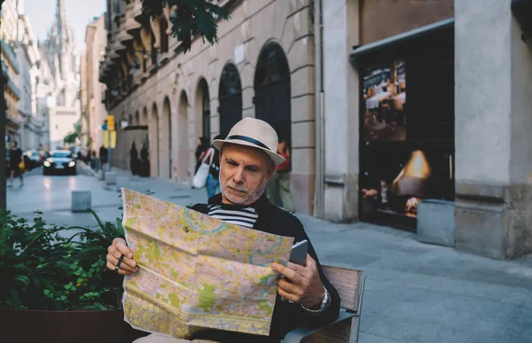 Aged male tourist reading orientation info on location map holding smartphone technology in hand and resting on city bench