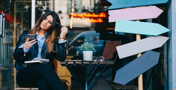 Genç bir kadın başını yana eğmiş bir şekilde, dizüstü bilgisayarlı ve akıllı telefonlu bir kafede otururken...