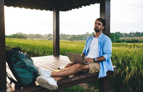 Kırsal kesimde dizüstü bilgisayarla uğraşırken ahşap çardakta uyuyan gündelik kıyafetli, yorgun genç girişimci.