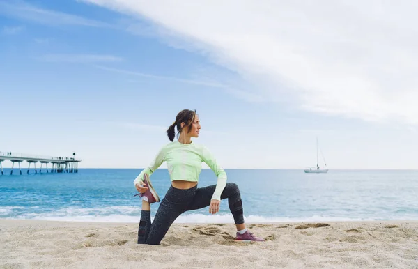 スリム40歳黒髪女性でアクティブウェアをしながらねじれでサポート脚と練習ヨガオン砂浜近く桟橋 — ストック写真