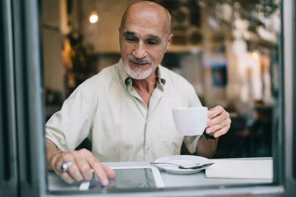 白种人老年男性用户在咖啡店通过数字平板电脑上网浏览网页信息的窗口视图 — 图库照片