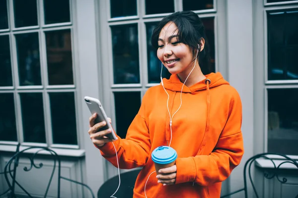 Delighted ethnic female in vivid hoodie standing on street with paper cup of takeaway coffee having video call on mobile phone