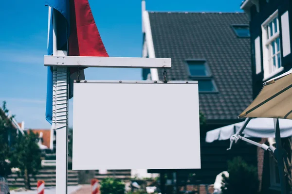 Blank ad sign with empty space near flag of Netherlands placed on street of contemporary town with building and street cafe