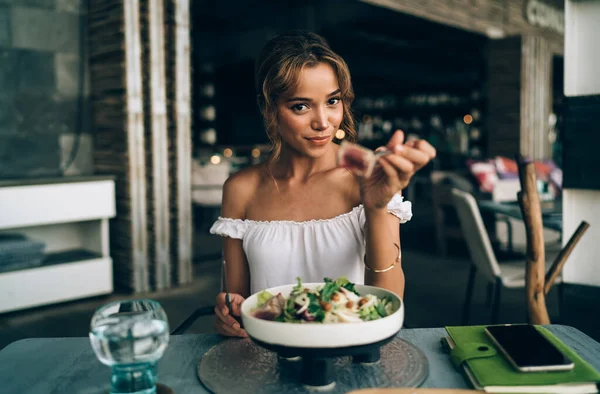 Young female wearing summer outfit and sitting in restaurant chair with bowl of healthy salad and glass of water