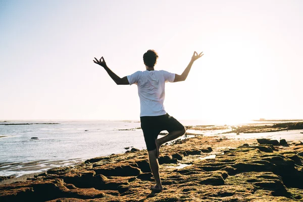 Back view full body unrecognizable male in casual summer outfit meditating while balancing on one leg with arms outstretched