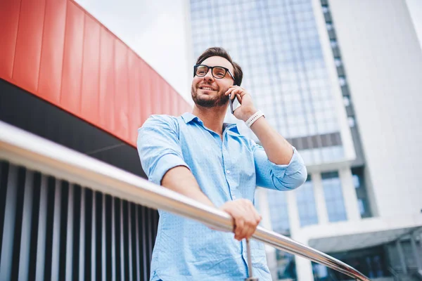 Smiling male tourist in eyeglasses satisfied with good international connection while talking on mobile with friend, handsome hipster guy enjoying phone conversation having cheap tariffs in roaming