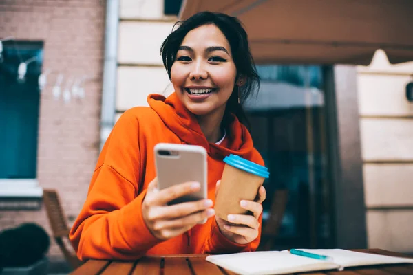 Smiling Asian female in vivid outfit looking at camera while sitting in street cafe with takeaway coffee and using smartphone for chatting