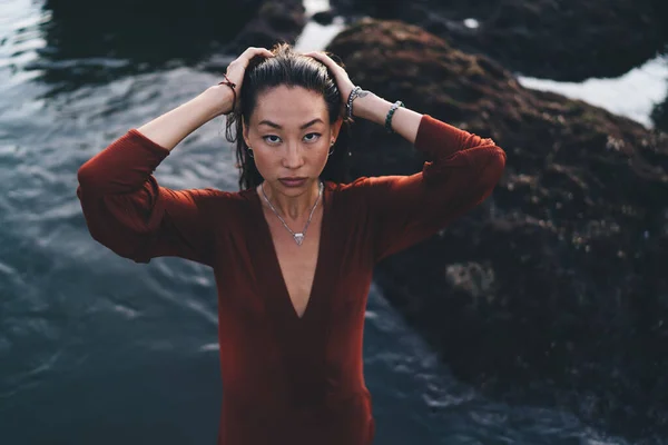 From above of confident female model with bracelets on hands and suspension on neck touching hair while looking at camera