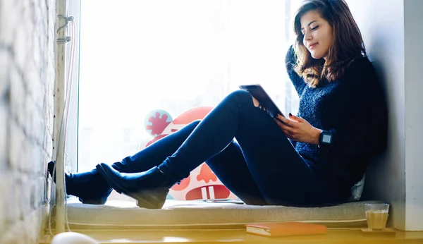 Side view of young remote worker checking notifications on tablet while sitting on cozy windowsill with coffee and notepad