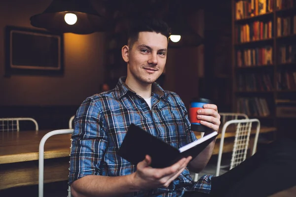 Cheerful young guy in blue shirt with paper cup of coffee and book sitting at library and smiling at camera