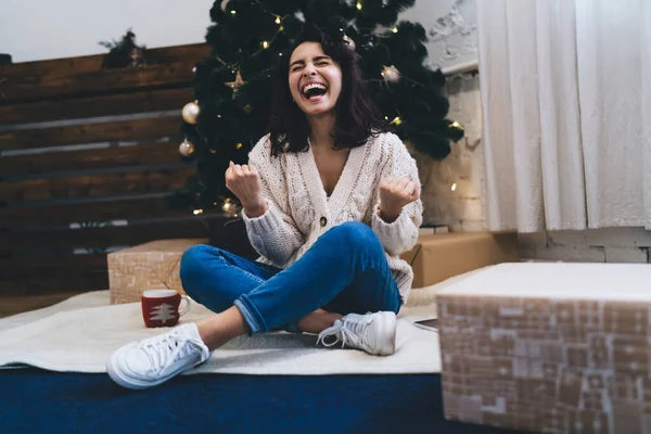 Full body of cheerful glad female screaming from joy among boxes with presents near Christmas tree with bright luminous garland