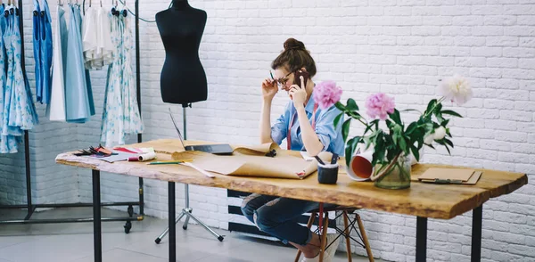 Düşünceli kadın terzinin ahşap masada oturan müşteriyle cep telefonuyla konuşmasının yan görüntüsü dizüstü bilgisayarı ve çiçekli vazosu.