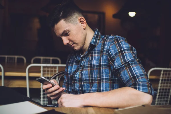 Mavi kareli gömlekli genç adam karanlık odada oturmuş akıllı telefonuyla internette sörf yapıyor.