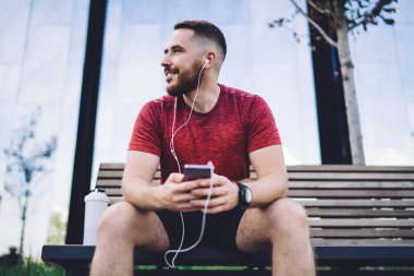 Kulaklıklı neşeli sakallı erkek antrenmandan sonra dinlenirken müzik dinliyor ve şehirdeki ahşap bankta otururken gözlerini kaçırıyor.