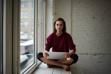 Sıradan giysiler içinde, pencerenin kenarında oturan çapraz bacaklı ve tabletle evde gezinen kameraya bakan konsantre kadın serbest yazar.