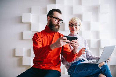 Sıradan giysiler içinde konsantre sakallı bir adam hafif modern çalışma alanında çalışırken dişlek tebessümle akıllı telefon ekranlı serbest çalışan bir kadın gösteriyor.