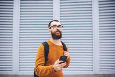 Sarı kazaklı ve gözlüklü genç adam. Sırt çantalı, cep telefonlu. Modern binanın yanında.