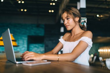 Beyaz bluzlu şık bir bayan rahat bir kafeteryada oturmuş gülümserken netbook klavyesiyle uzaktan çalışıyor.