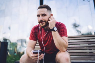 Genç sakallı erkek spor kıyafetler giyiyor. Cep telefonu kullanıyor. Şehir sokağındaki ahşap bankta dinleniyor ve müziğin keyfini çıkarıyor.