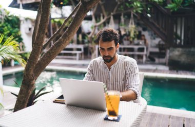 Yazın ağaç ve havuz başında internette internette gezinen sakallı, dalgın, yakışıklı genç bir erkek.