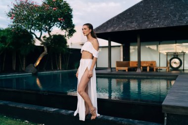 Havuz kenarındaki ahşap terasta tek başına dinlenirken şık giyinmiş ve gözlerini kaçıran çıplak ayaklı turistin yan görüntüsü.