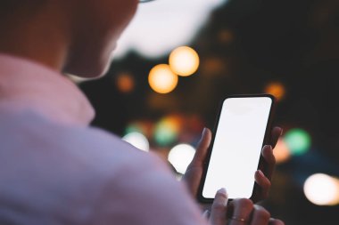 Gözlüklü tanınmayan şahıs cep telefonuyla sosyal medyayı kontrol ediyor ve arka planda Bokeh ışıkları ile şehir caddesinde dururken internet kullanıyor.