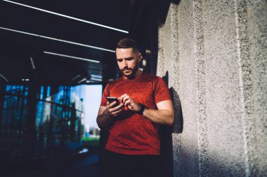 Genç sakallı erkek, güneşli bir günde çatıda dikilirken duvara yaslanırken cep telefonunun ekranına dokunuyor.