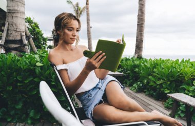 Terasta sandalyede deniz kenarında oturan ve tatillerini geçirirken tablette roman okuyan gündelik giysiler içinde çekici bir kadın vücudu.