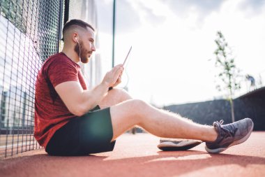 Spor ayakkabıları ve tablet tutan erkek çalıştırıcının zemin seviyesi sokak egzersizlerinden sonra dinlenirken