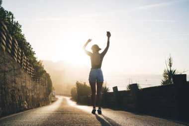 Gündelik giyimli, mutlu, beyaz tenli bir kadının arka görünüşü. El kaldırıyor, yaz tatillerinden zevk alıyor.
