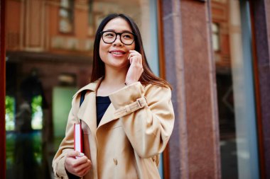 İyimser Asyalı kız öğrenci, dişlek tebessümle elinde defter, elinde cep telefonuyla gezerken başka tarafa bakıyor.