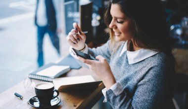 Tebessümlü, dişlek bir bayan öğrencinin elinde kahve ve not defteriyle masada otururken akıllı telefonlara göz gezdirirken