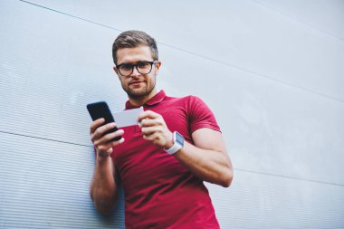 Sakallı, sıradan kıyafetleri ve kol saati takmış elinde kartvizitle bekleyen ve gri duvarda akıllı telefonları karıştıran sakin bir adamdan.