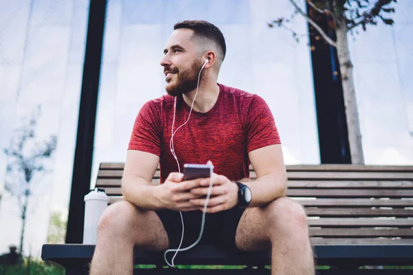 Cheerful bearded male in earphones listening to music while resting after training and looking away while sitting on wooden bench in city